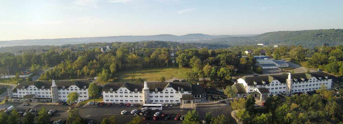 Aerial view of a large white hotel with dark roofs, surrounded by trees and a parking lot.