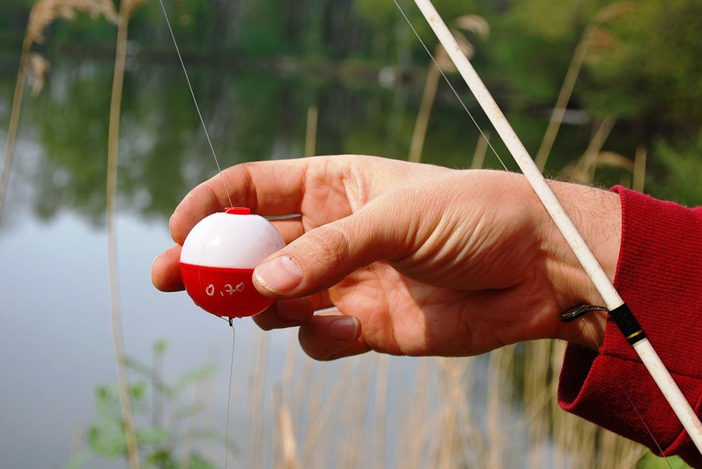 A mans hand holds a red and white fishing bobber in front of the lake.
