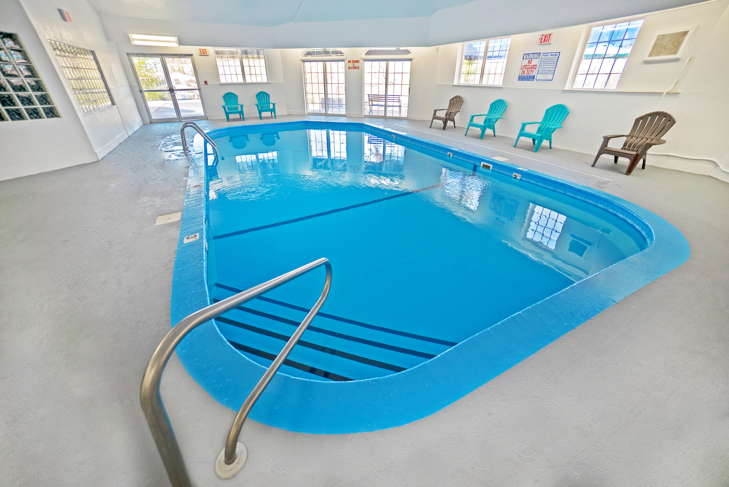 An indoor pool with blue water and a metal railing, surrounded by chairs and windows.