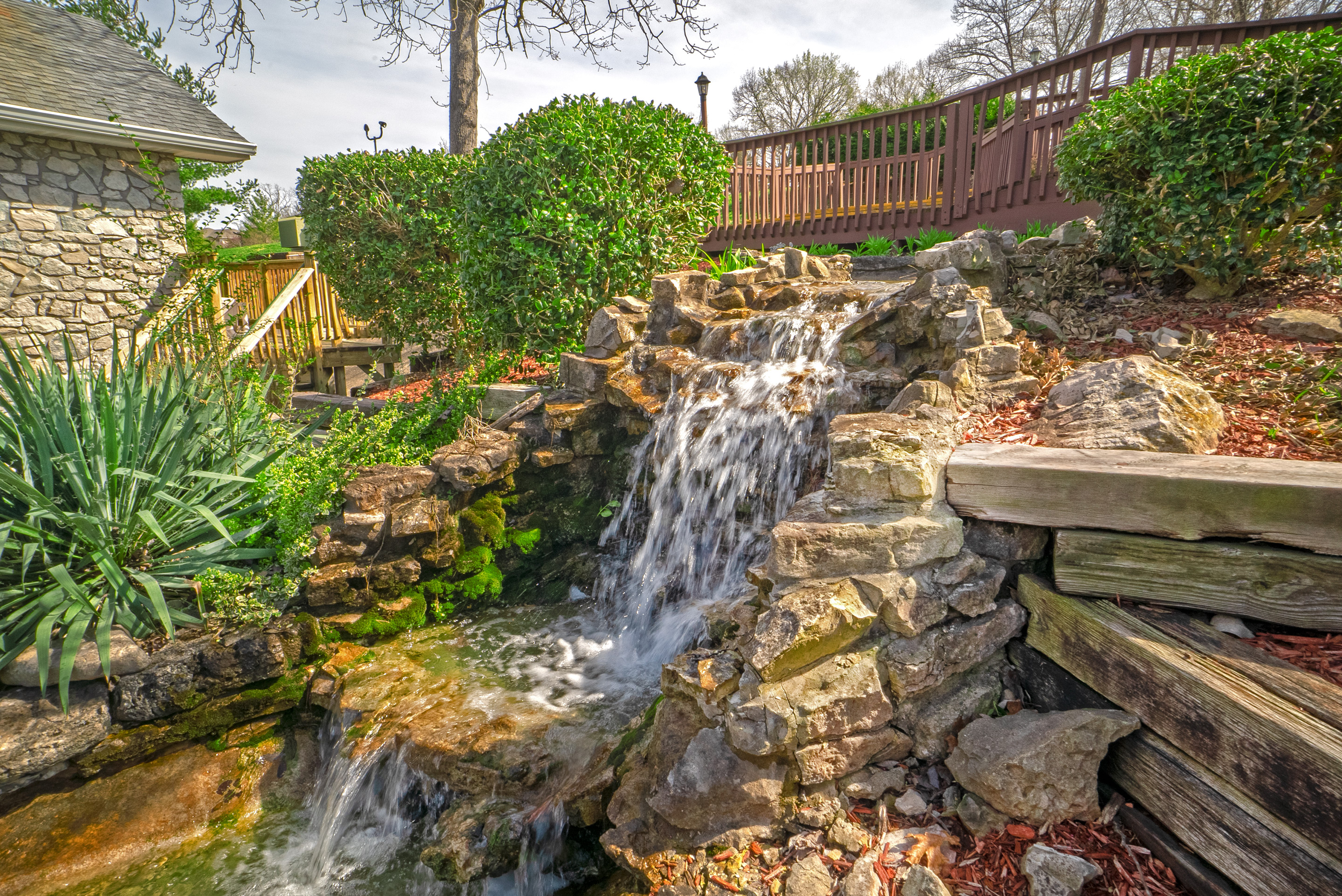 A small waterfall cascades down a rocky slope, surrounded by lush greenery and a wooden bridge in the background.