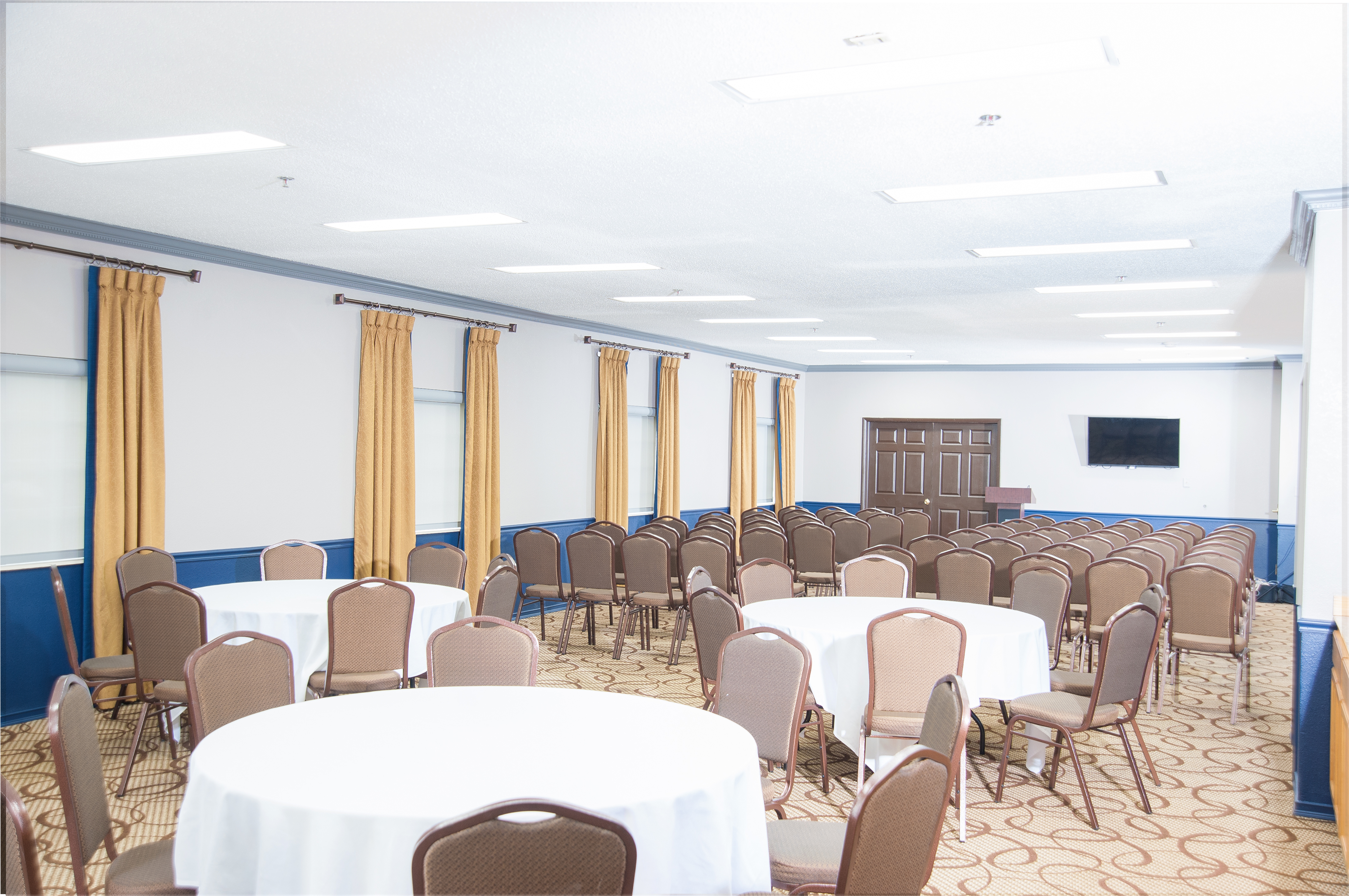 A large empty meeting room with round tables, chairs, a TV, and yellow curtains.