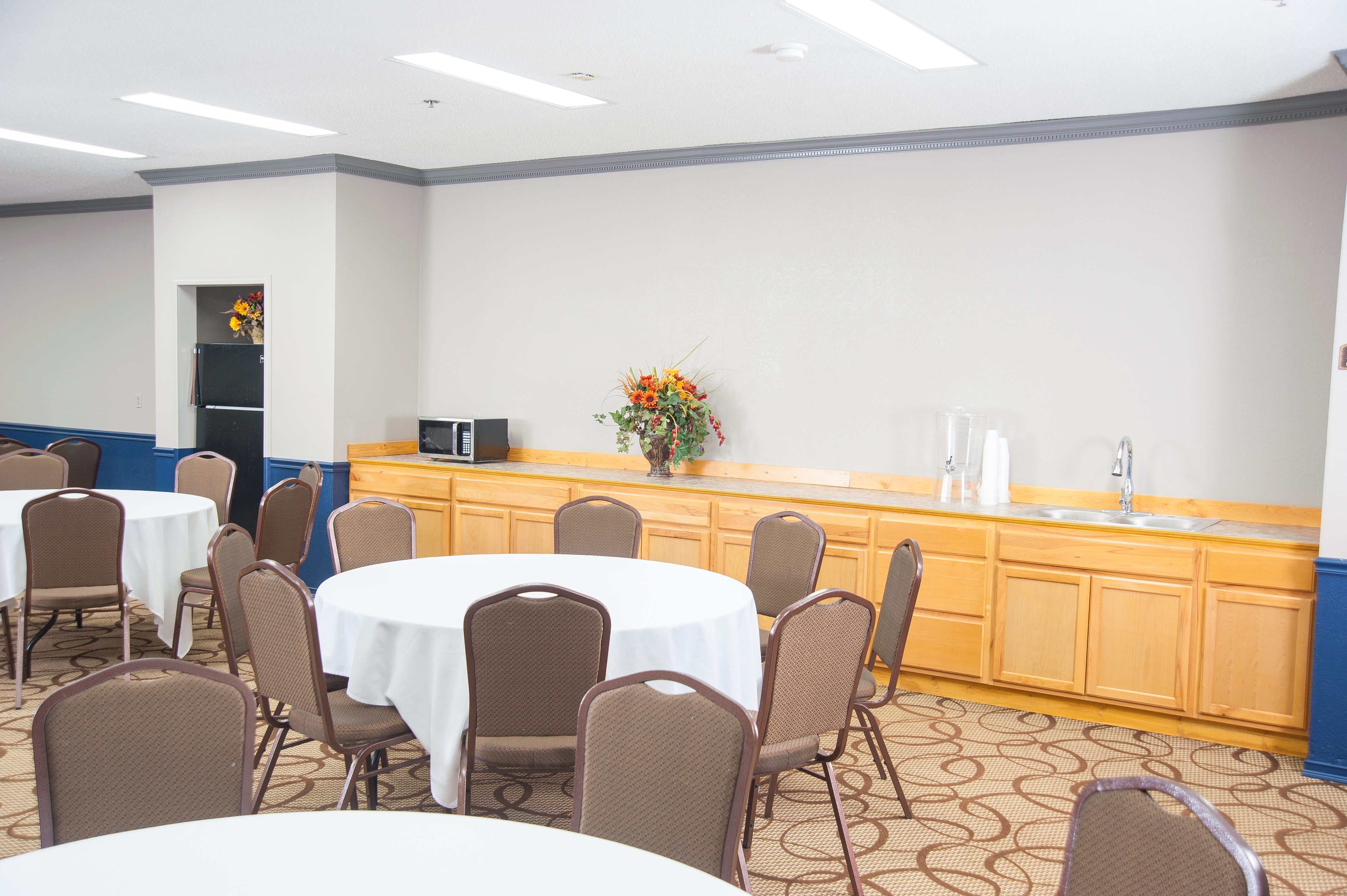 A meeting room with round tables, brown chairs, wooden cabinets, and a sink.